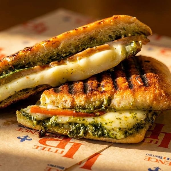 Two halves of a grilled panini with melted cheese, pesto, and tomato on branded paper.